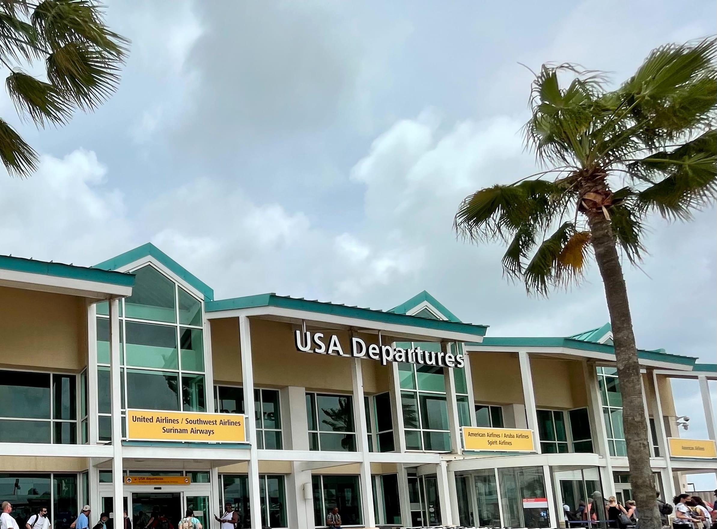 The USA Departures building at Queen Beatrix International Airport, Aruba, with airline signs over the doors and palms in the wind