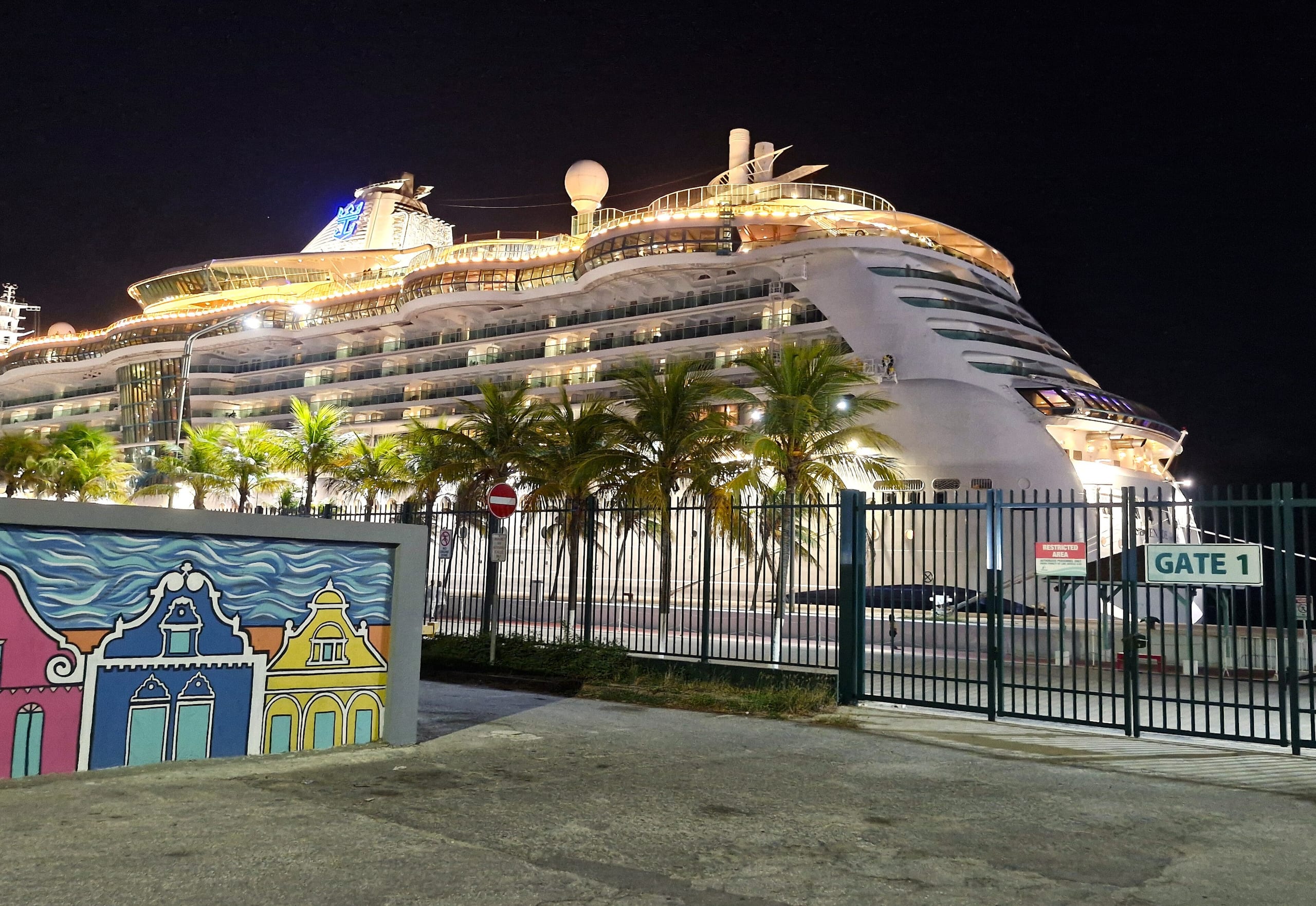 A lit cruise ship docked behind the Gate 1 fence at the Oranjestad cruise terminal, Aruba, at night, beside a mural of painted gabled houses