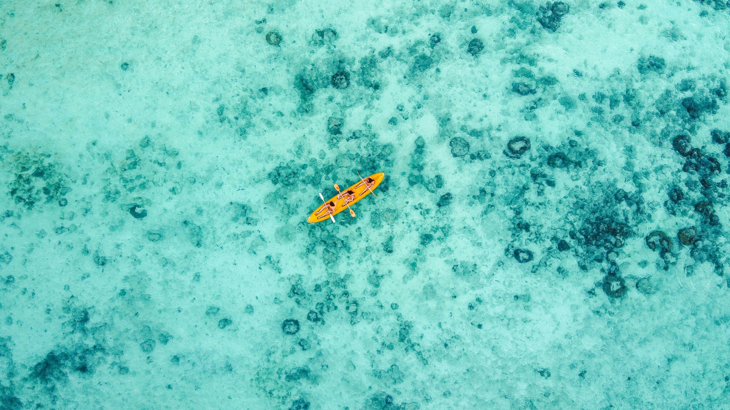 An orange kayak on pale turquoise water at El Nido, seen from directly above with dark coral patches scattered under the surface
