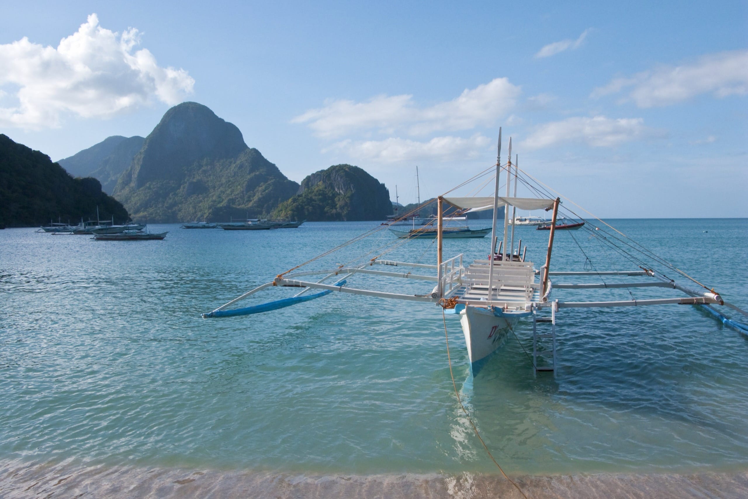 Empty outrigger boat tied in shallow turquoise water with limestone hills and moored boats behind it