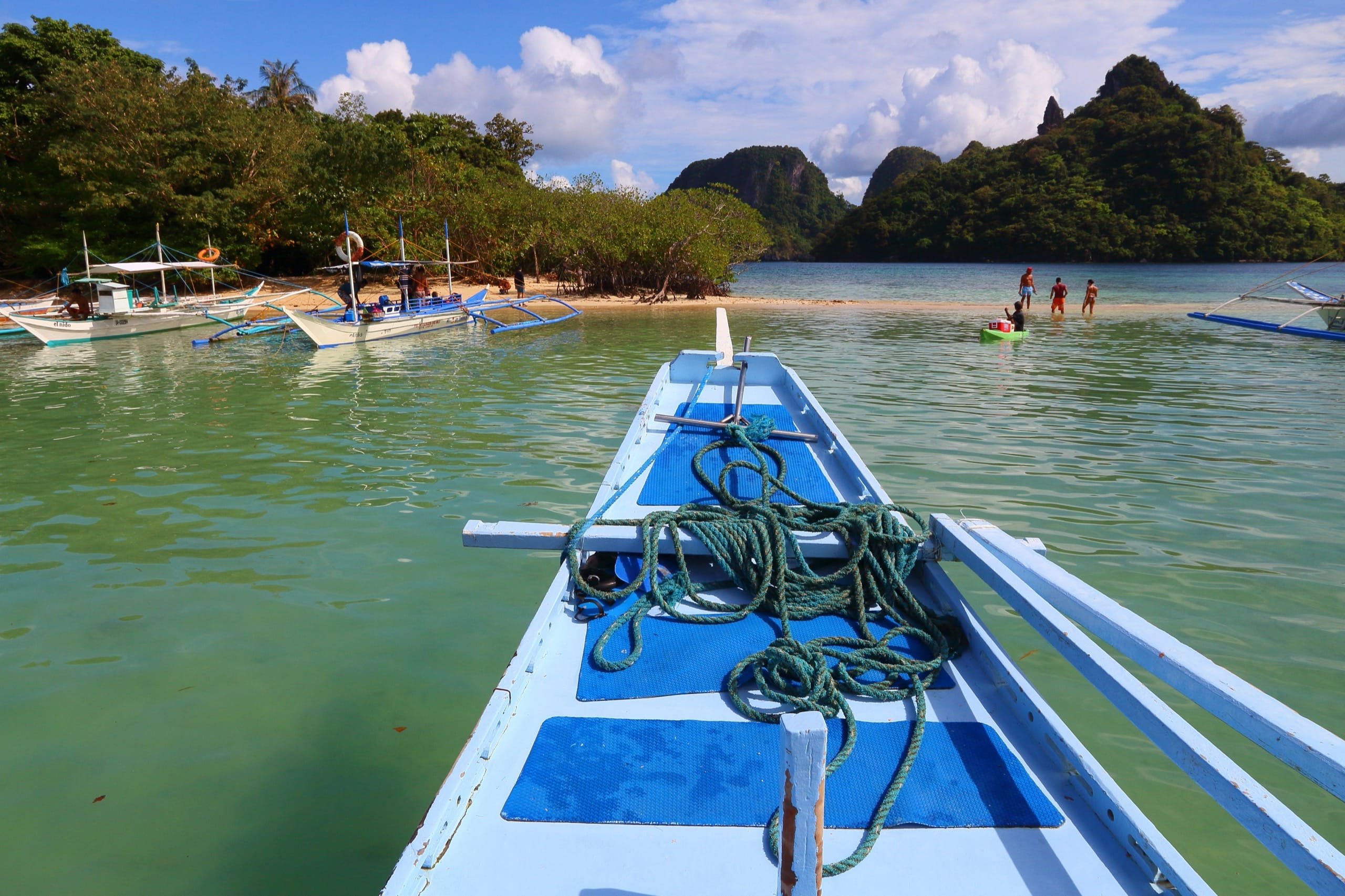 Outrigger boat bow in green water at a sandbar with moored boats and forested limestone hills beyond