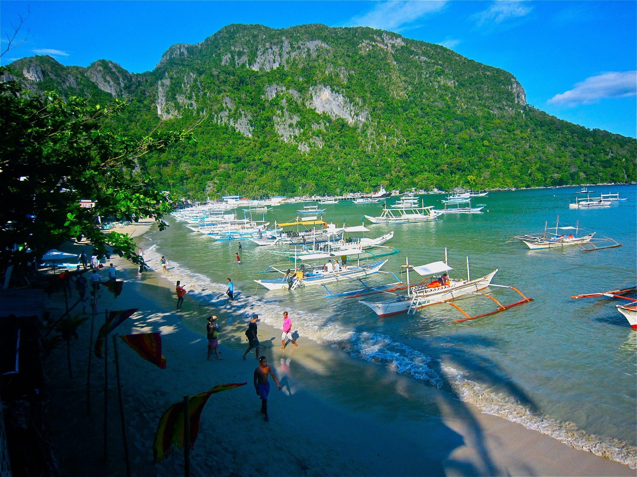Rows of outrigger tour boats waiting along a beach below a forested limestone hill, people wading between them