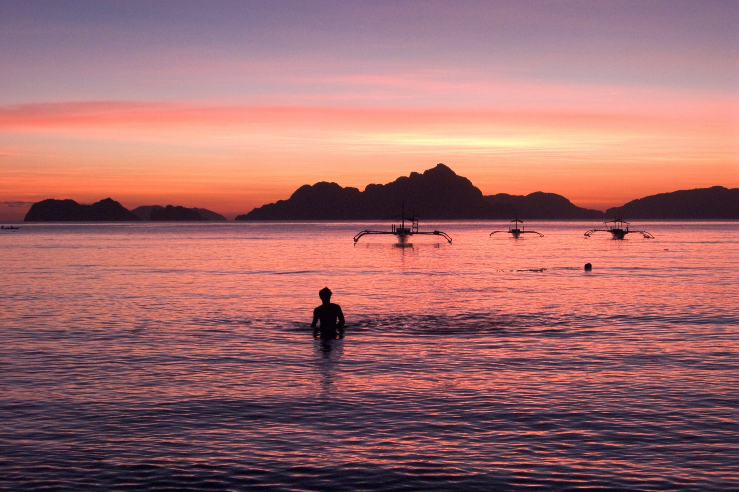 Silhouetted swimmer and three outrigger boats against a pink and orange sunset over the Bacuit Bay islands