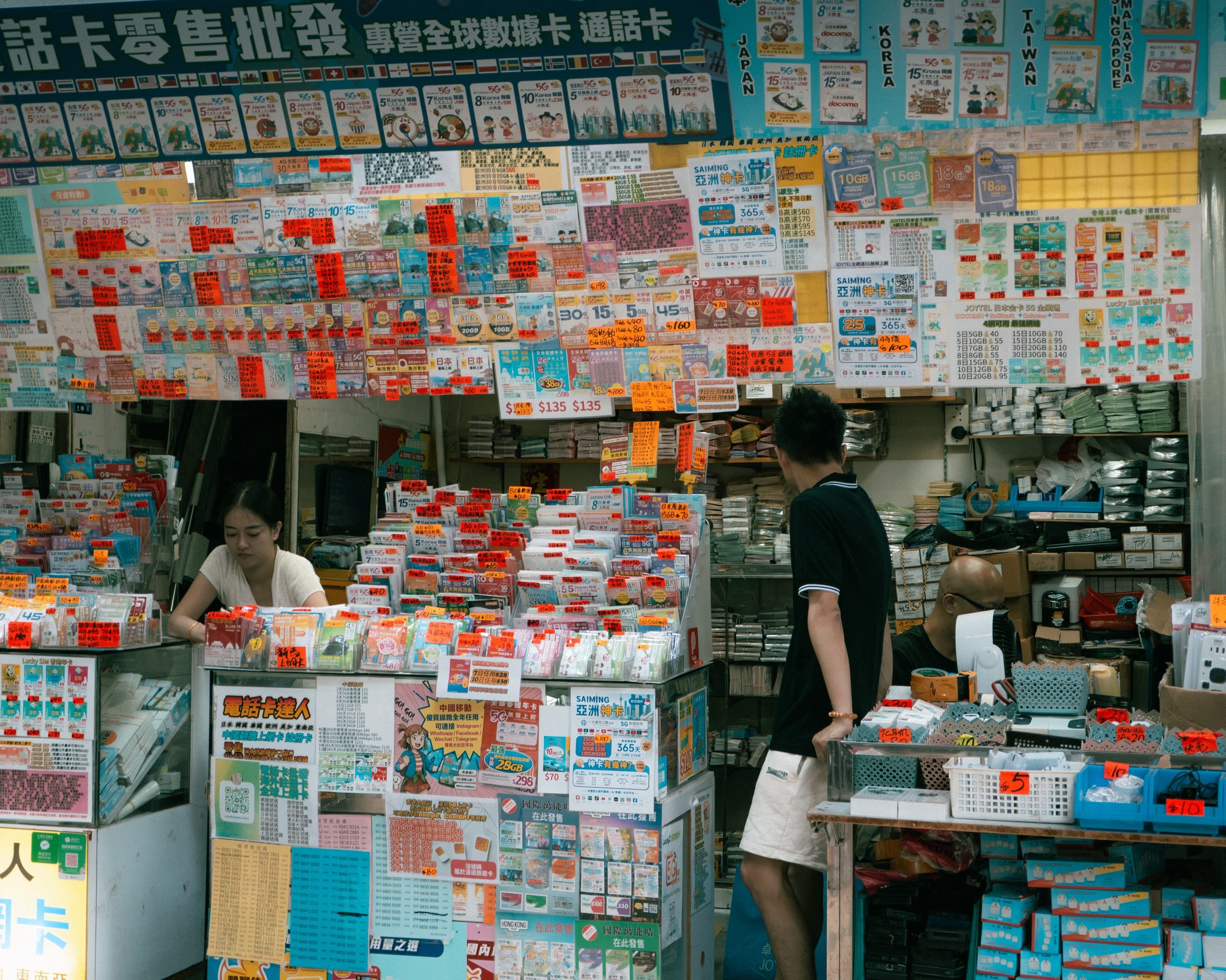 A phone card stall in Kowloon, Hong Kong, its walls covered in prepaid SIM packs and price tags, a customer at the counter