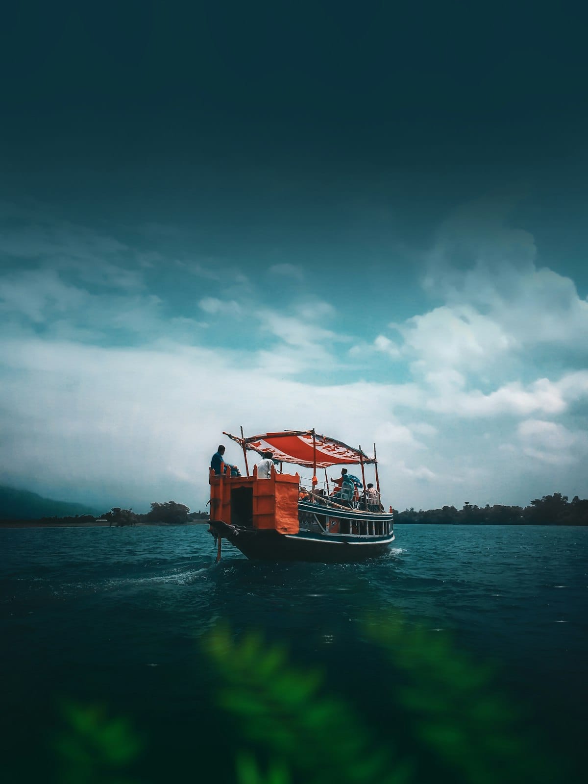 A wooden passenger boat under an orange canopy crossing dark green water, a wooded shoreline and heavy cloud behind it