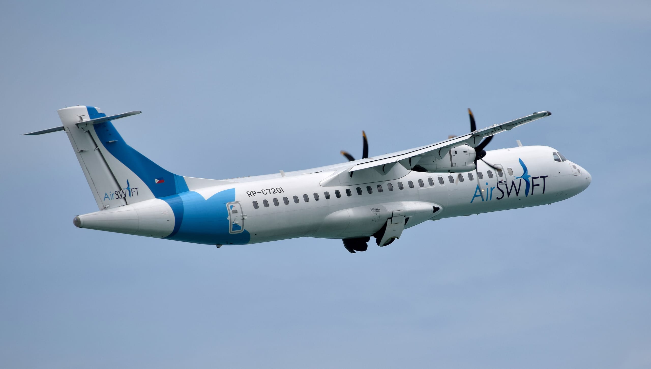 An AirSWIFT turboprop climbing out against a pale blue sky, undercarriage still coming up