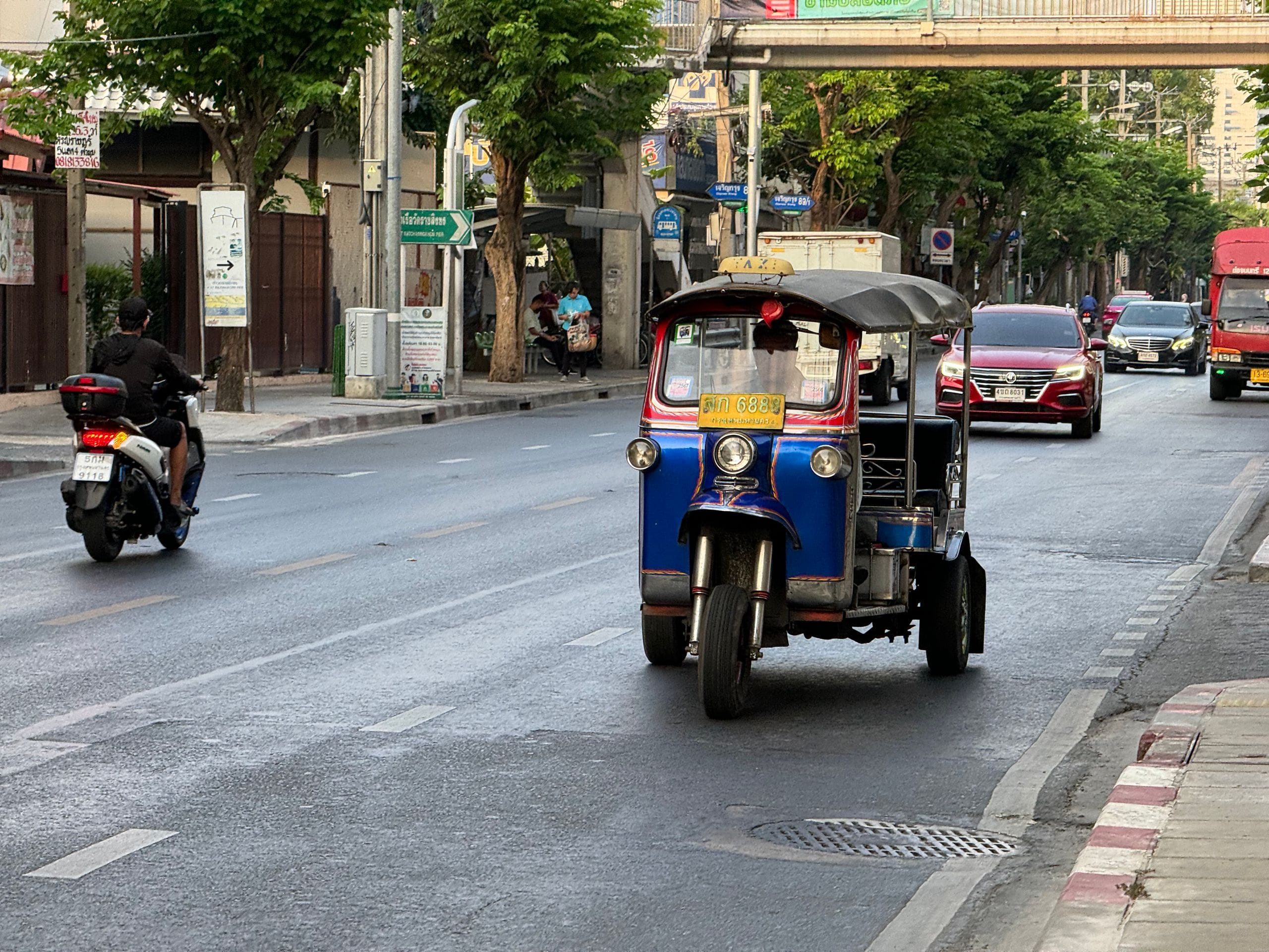 A blue and red three-wheeled tuk-tuk driving along a tree-lined city street beside a scooter