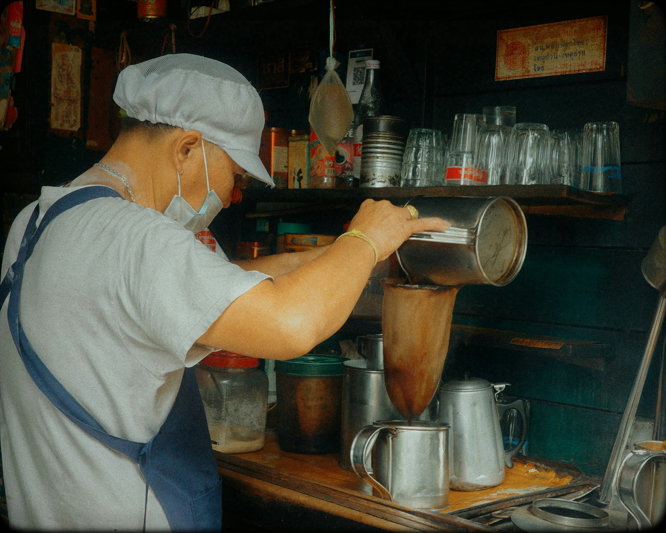 A vendor in a mesh cap pouring hot coffee through a cloth filter at a street counter