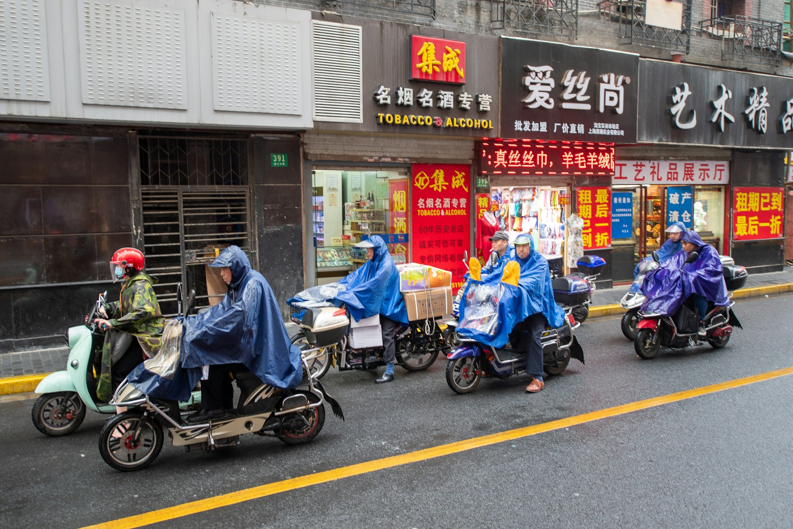 Scooter riders in blue rain ponchos lined up on a wet shop-lined street