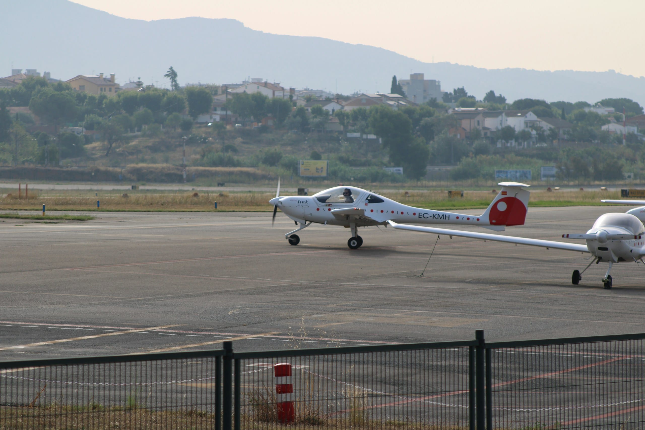 A small propeller plane taxiing on the apron of a small airfield with hills behind