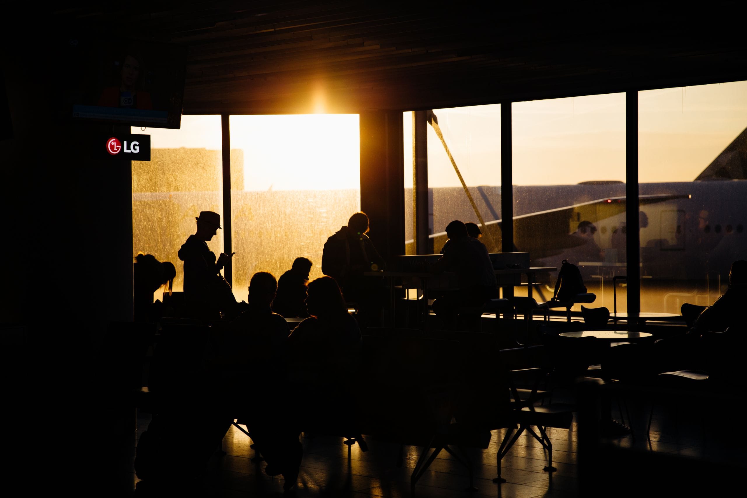 Silhouetted travelers waiting in a terminal against a low gold sun and a parked airplane