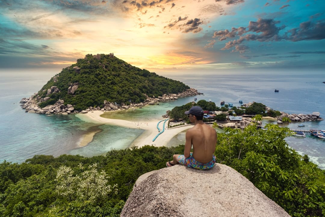 A person sitting on a boulder above Koh Nang Yuan off Koh Tao in Thailand, two islands joined by a sandbar with boats moored in the shallows below