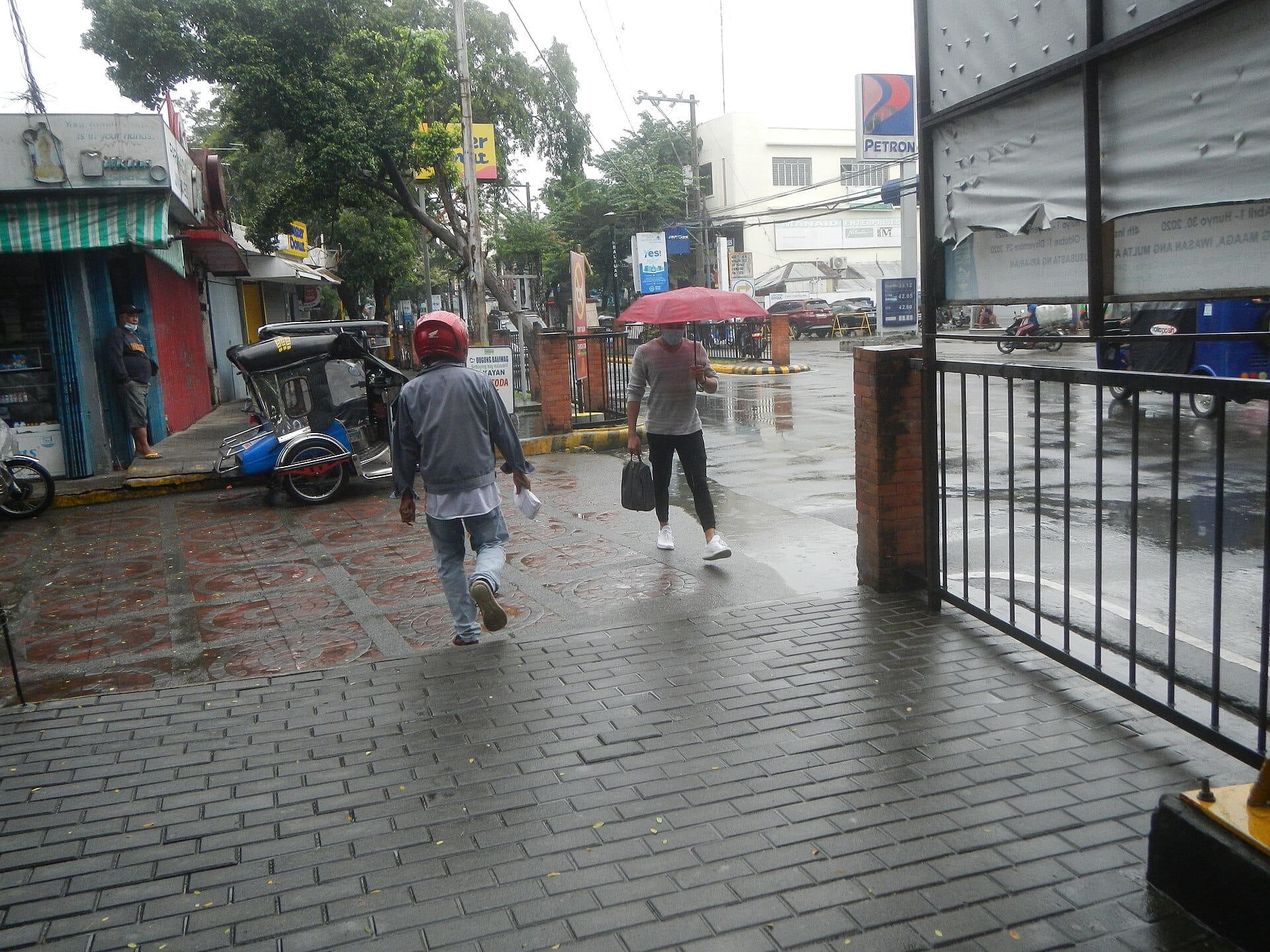 Pedestrian with a red umbrella crossing a wet paved street beside a parked tricycle in Baliuag, Bulacan