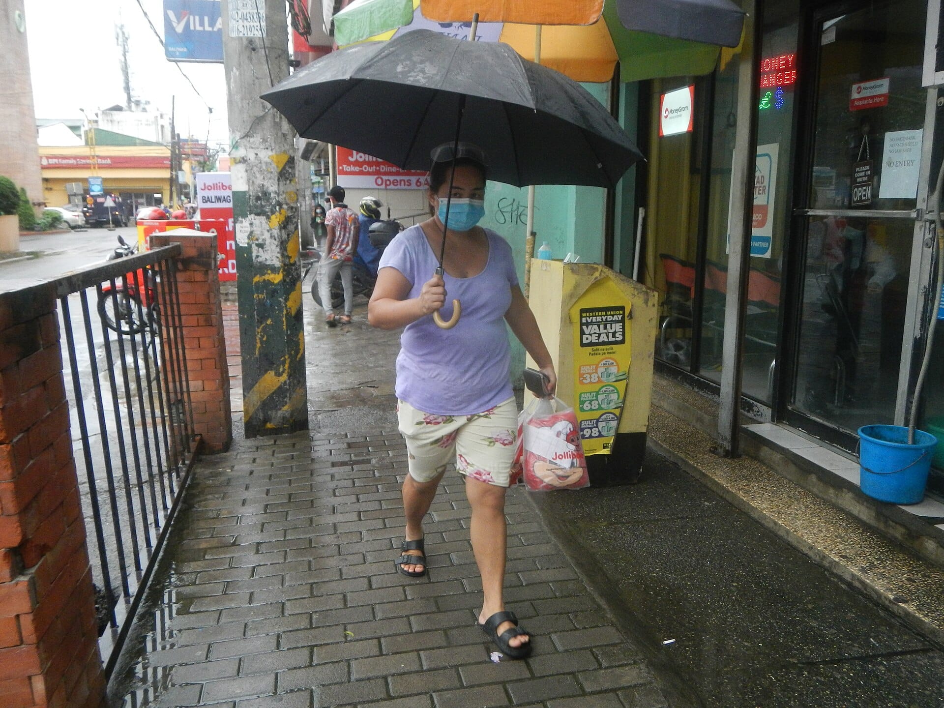 Woman in a face mask carrying a shopping bag under a black umbrella on a wet sidewalk in Baliuag, Bulacan
