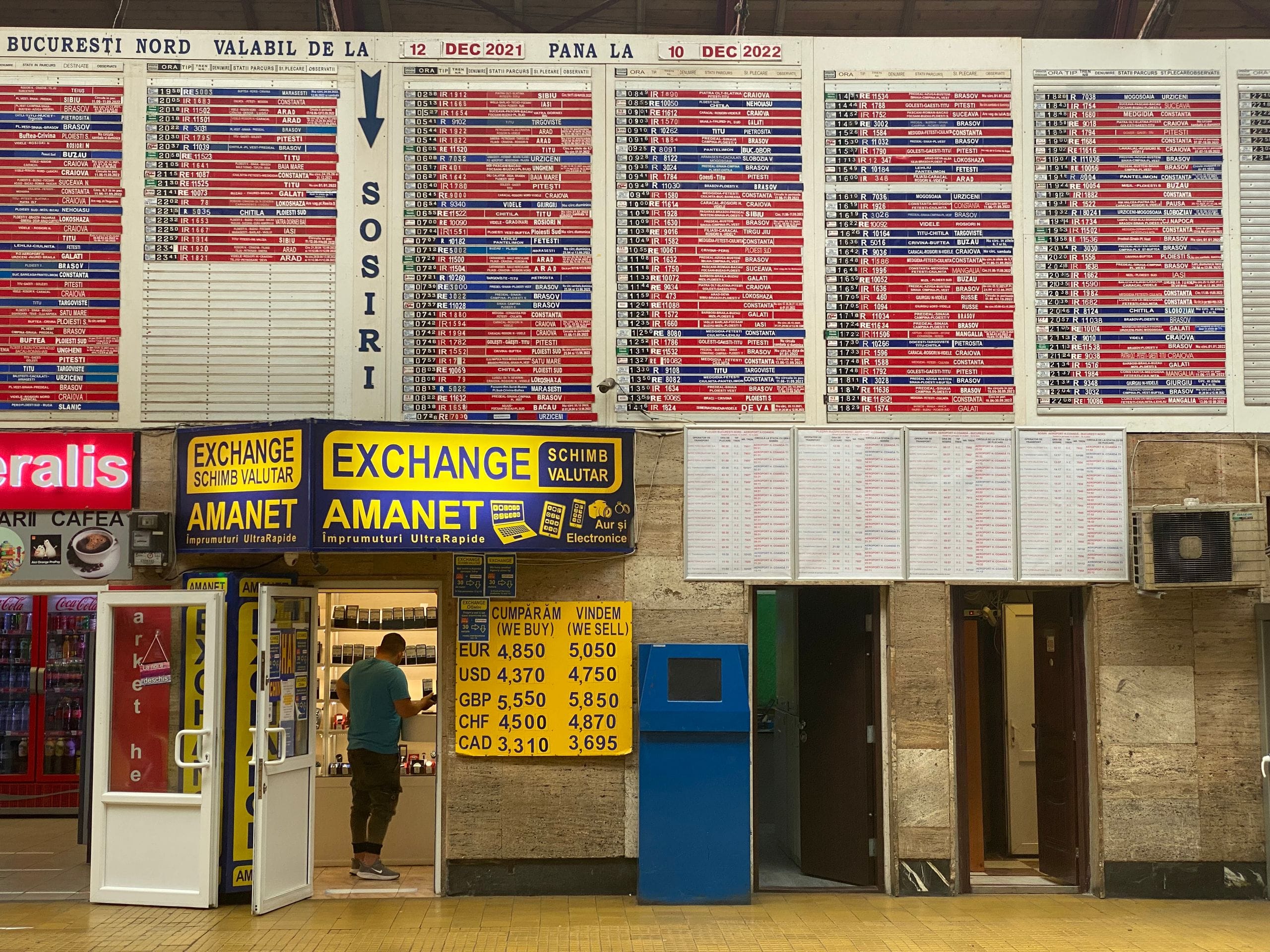 A yellow currency exchange board listing buy and sell rates beside a kiosk at Bucharest Nord station
