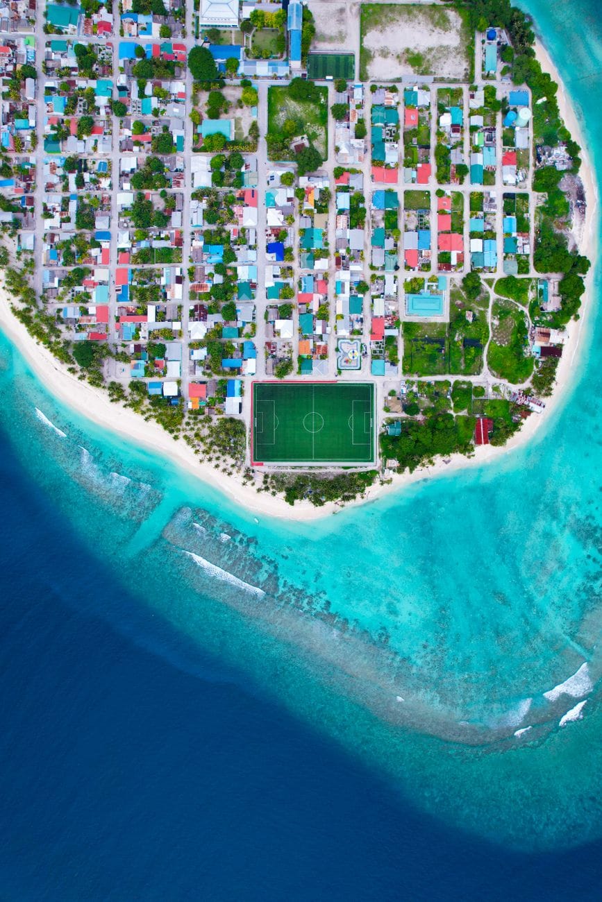 The inhabited island of Meedhoo in Raa Atoll from the air, its colored roofs packed in a grid beside a green football pitch above the reef