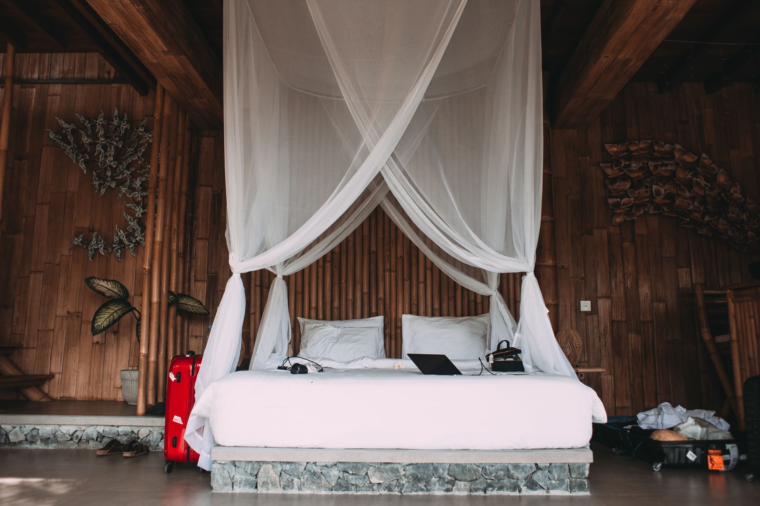 A double bed under a white mosquito net in a bamboo-walled room, a red suitcase and open luggage beside it