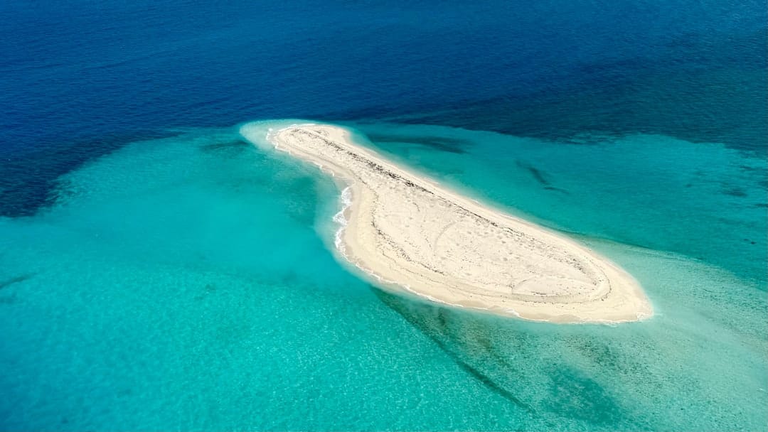 A bare sandbar surrounded by turquoise water fading into deep blue, seen from above