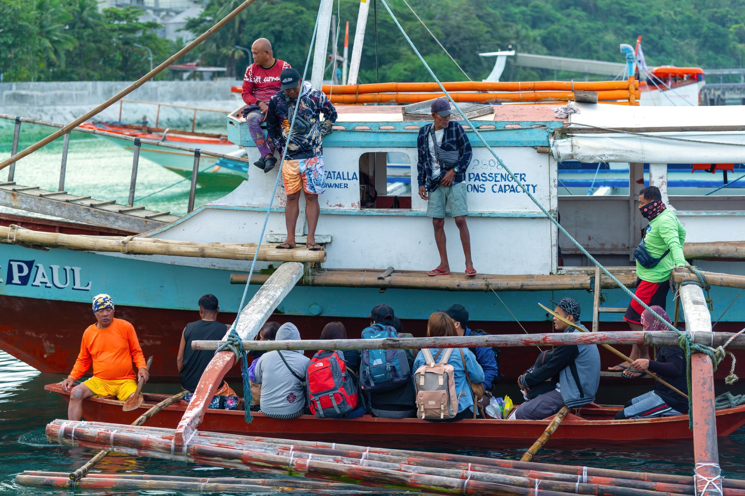 Passengers with backpacks sitting in a small outrigger boat beside a larger passenger boat, crew standing on deck