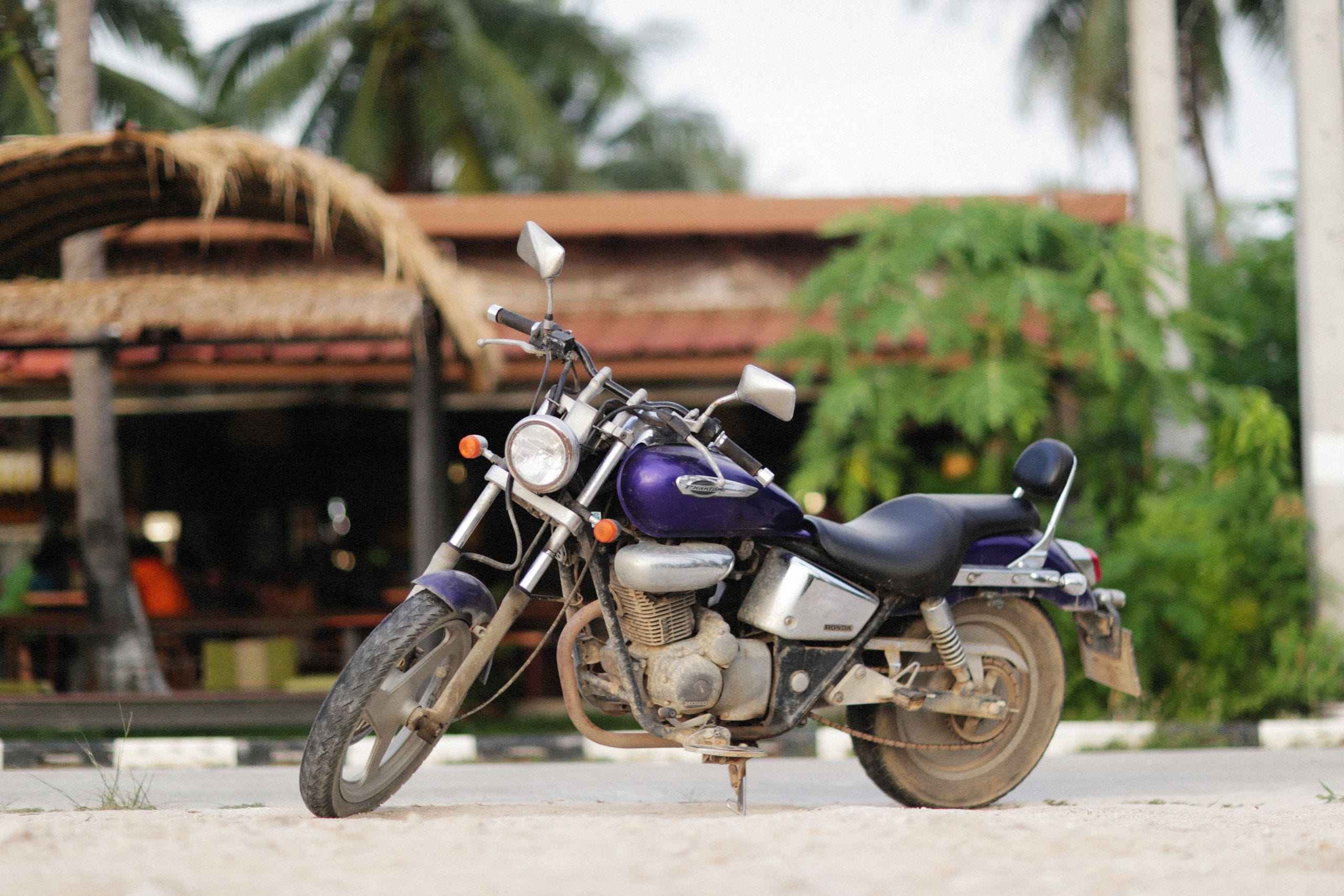 A purple motorcycle parked on a sandy lot in front of a thatched open-air cafe with palm trees behind