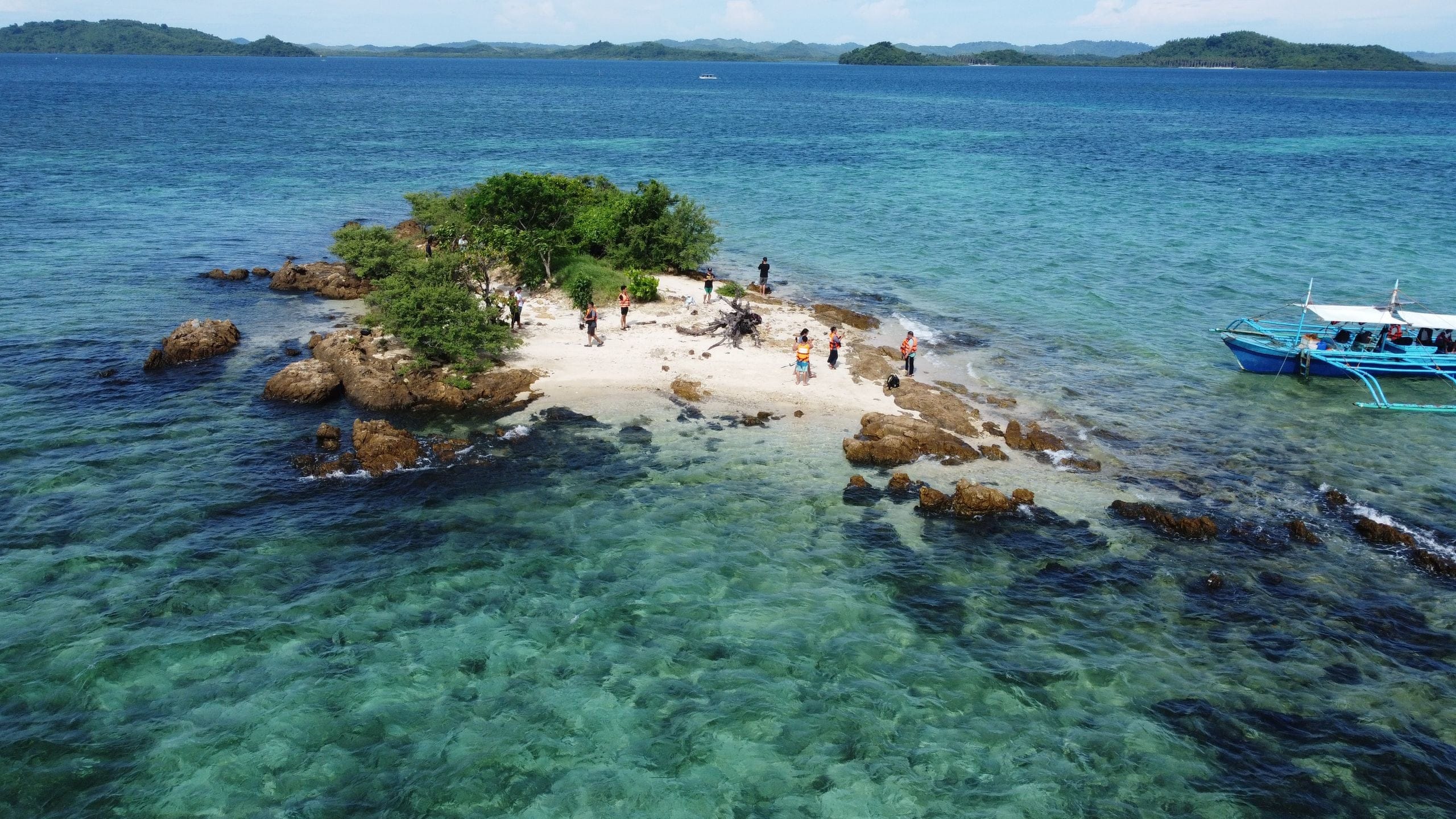 A small rocky islet off Dumaran in Palawan seen from the air, a few people on its sand and an outrigger anchored in the clear water beside it