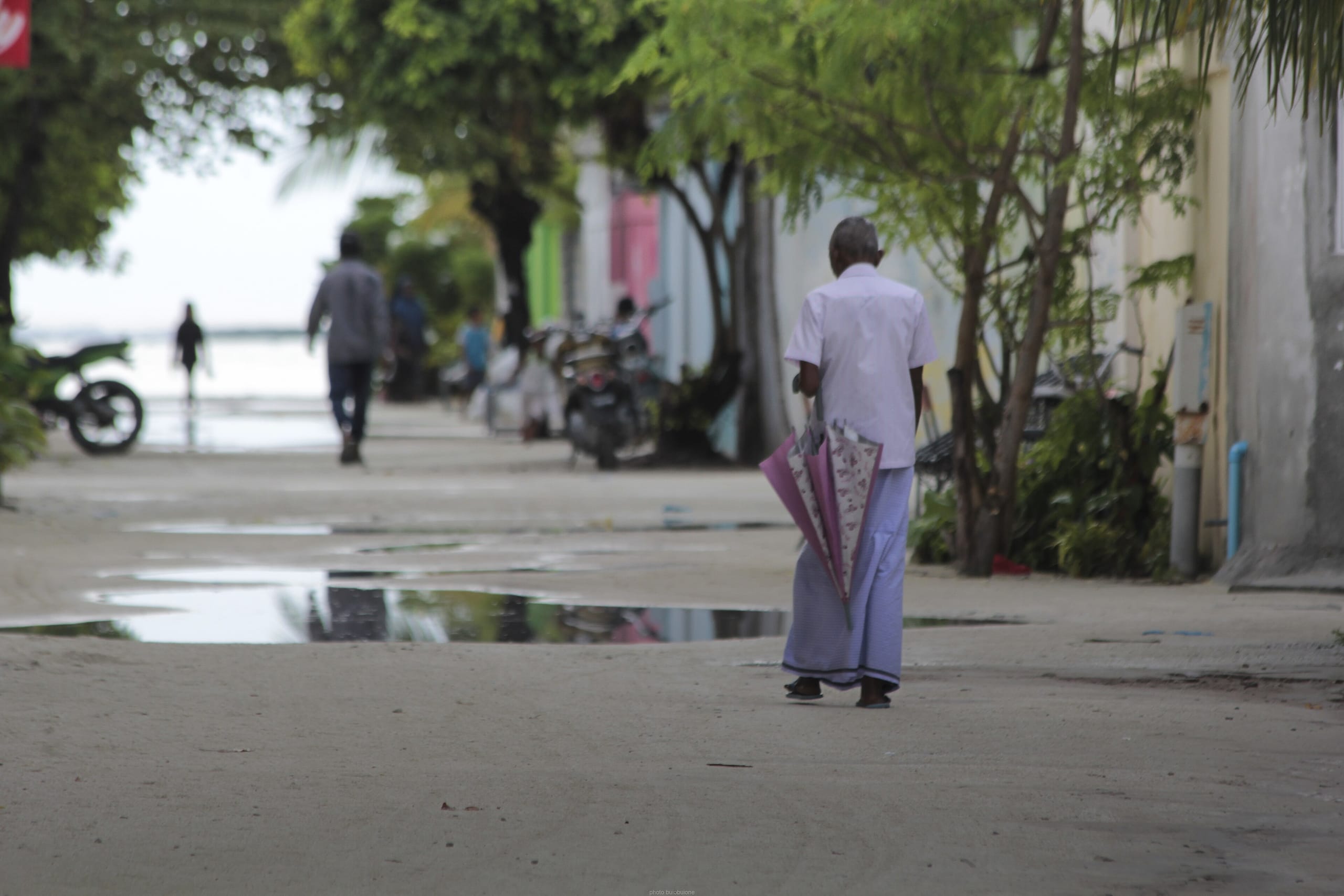 A man in a white shirt and sarong walking down a sandy street on Maafushi with a folded umbrella