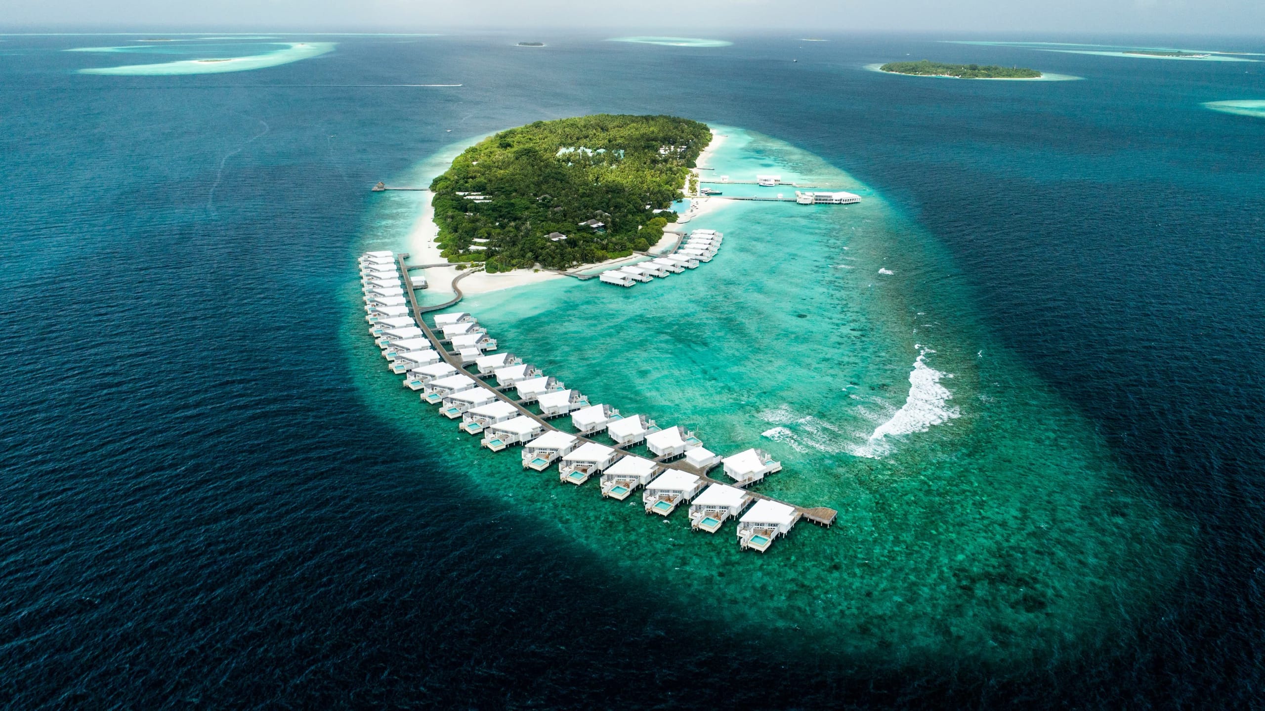 An oval island ringed by a continuous line of white overwater villas in deep blue water