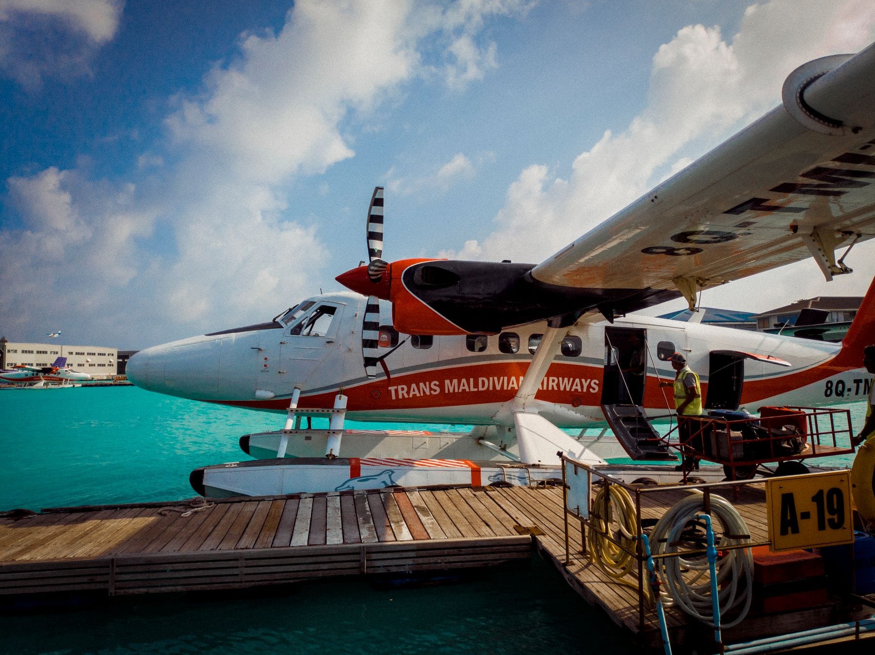A Trans Maldivian Airways seaplane tied to a wooden dock at the seaplane terminal, a handler loading bags through the open door