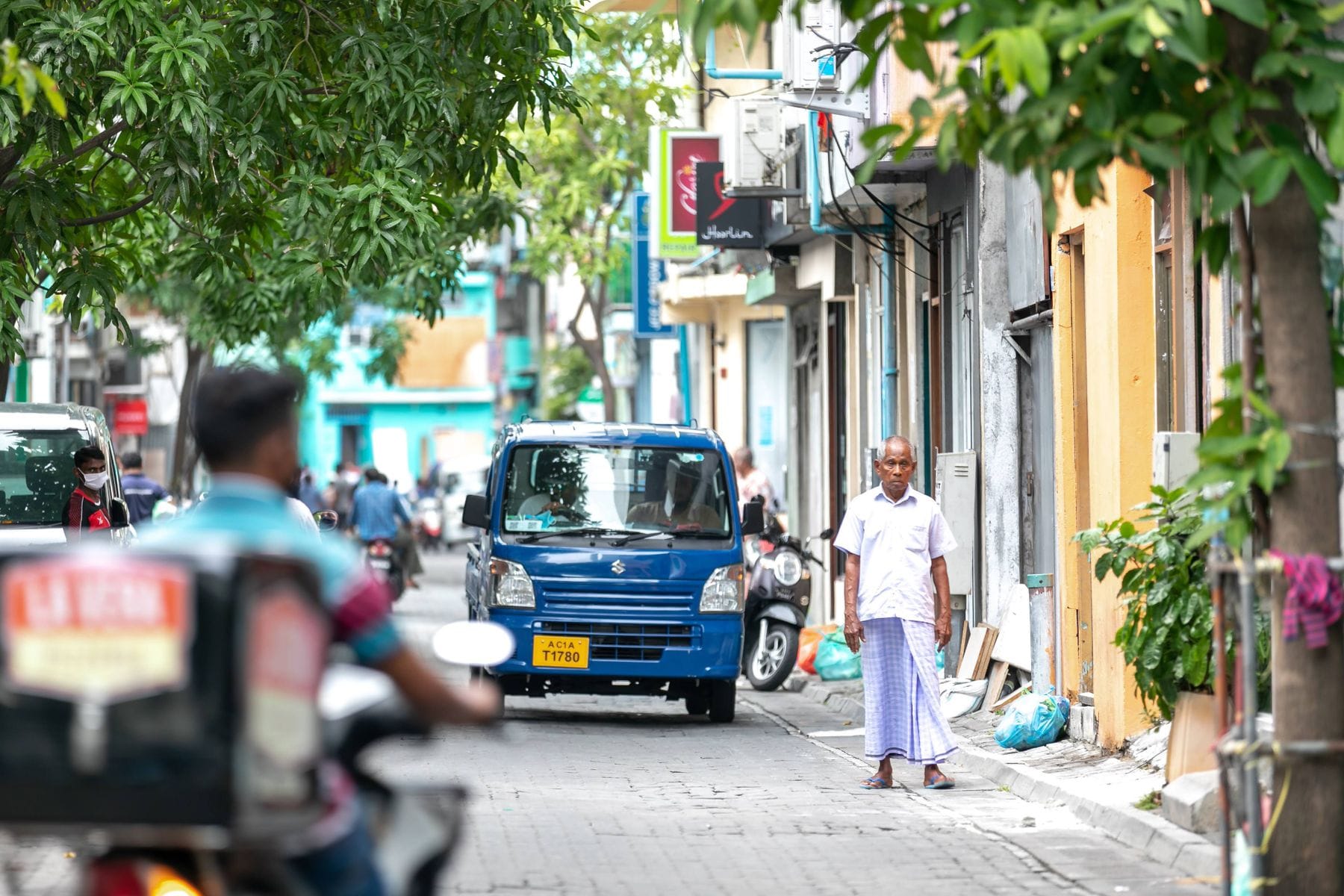 A street of shops in Malé with a small blue pickup, parked scooters and a man in a sarong standing on the curb