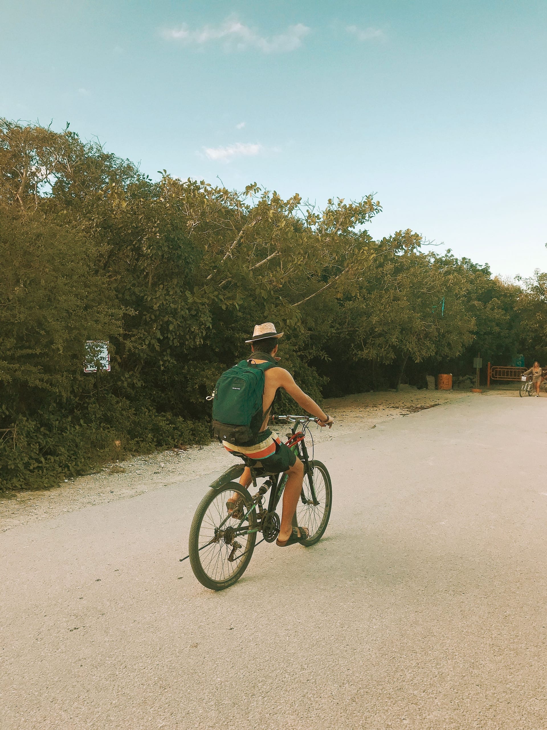 A man in a hat riding a bicycle away down a sandy track with a backpack on